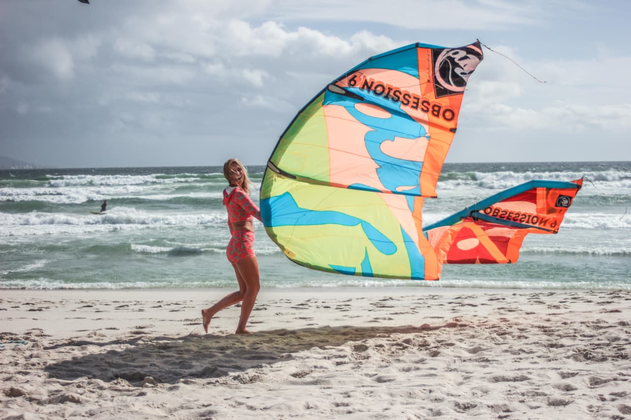 A kitesurfer walking down the Bloubergstrand beach with a bright kite, waves breaking behind