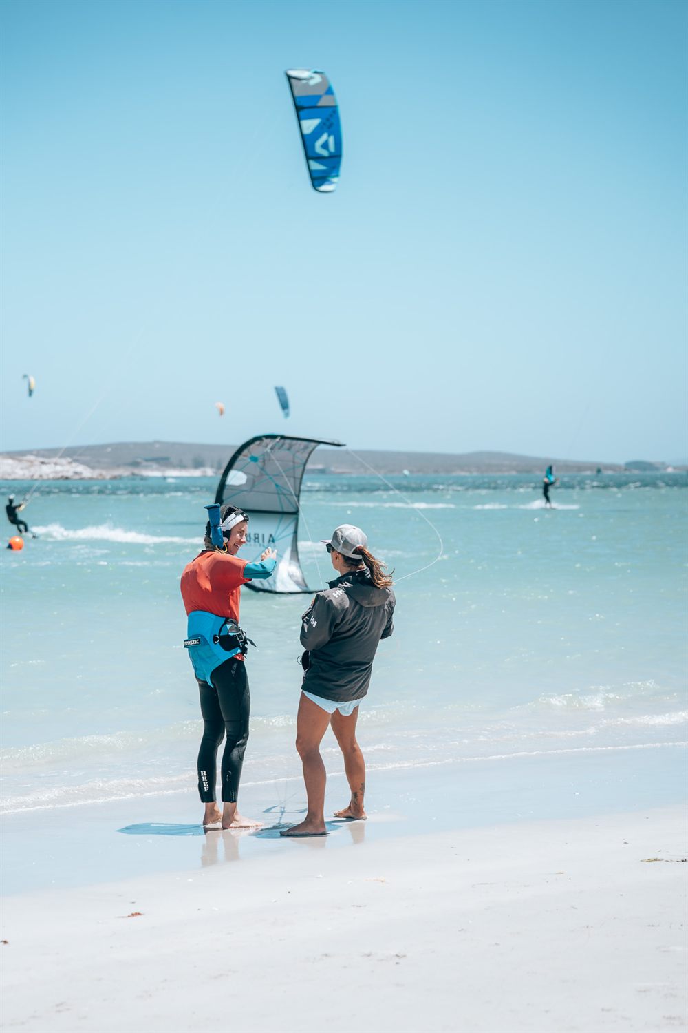 A kite instructor coaching a beginner on the beach at the water's edge, kites flying over the flat water behind them